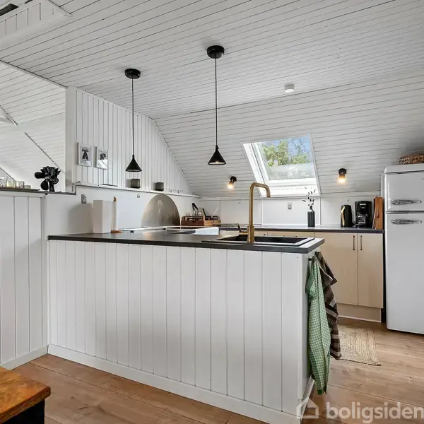 Et hvidt køkken med en sort bordplade har hængende lamper og moderne apparater som en retro køleskab. Store vinduer og skråloft giver lys og rummelighed.
