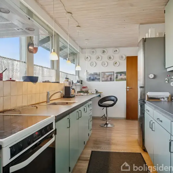 Køkken med mintgrønne skabe og sorte bordplader. Kaffemaskine ved siden af vask. Vægge dekoreret med tallerkener, billeder. Vinduer giver udsigt til blå himmel. Fliser bag komfur. Skammel ved hjørne.