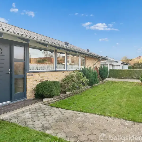 En dør med tallet 23 fører ind i et hus med murstensfacade. Huset ligger i en have med græsplæne og buske under en klar, blå himmel.