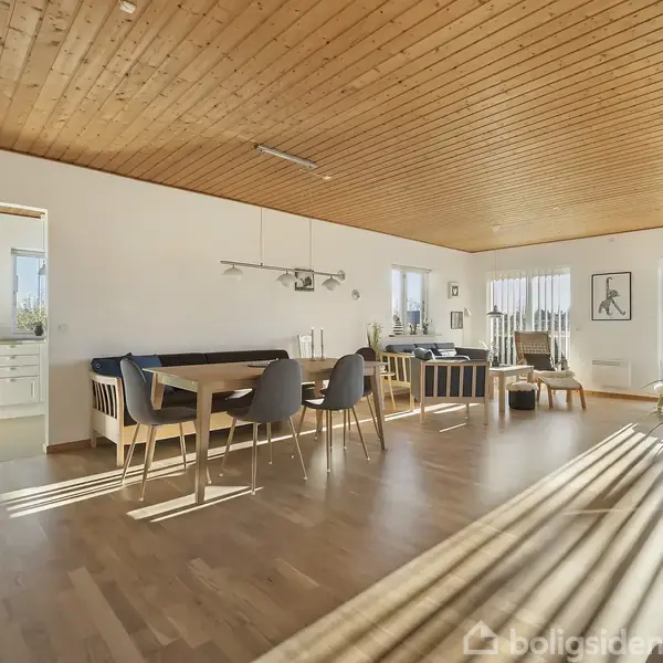 Stue med spisebord og stole, hvor sollys strømmer ind gennem store vinduer. Rummets trægulv og loft skaber en varm atmosfære. I baggrunden ses en åbning til køkkenet.