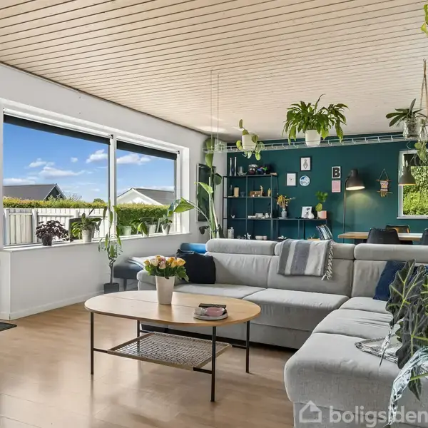 Stue med stor, grå hjørnesofa og runde sofaborde. Hylder med bøger og planter hænger på en blålig væg. Store vinduer lysner rummet, der viser udsigt til terrasse og have.