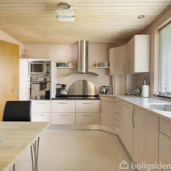 Et moderne køkken med lyse træskabe og stålapparater, herunder en ovn og emhætte. Der er en stor vask ved vinduet, et spisebord med stole, og rolig belysning.