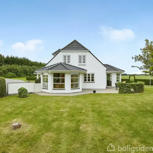Et hvidt hus med gråt tag står på en velplejet græsplæne. Der er hække og træer i baggrunden og en klar blå himmel.