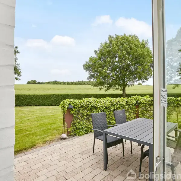 Havebord med sorte stole står på en terrasse med flisebelægning. En velplejet hæk omkranser græsplænen, og en enkelt træ giver skygge i baggrunden mod en grøn mark.