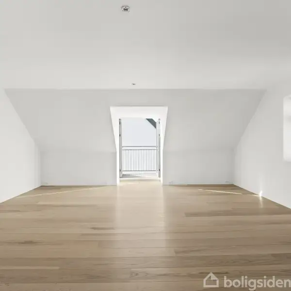 Et tomt rum med trægulv og hvide vægge leder op til en lille altan gennem en kviståbning; til højre ses et køleskab.