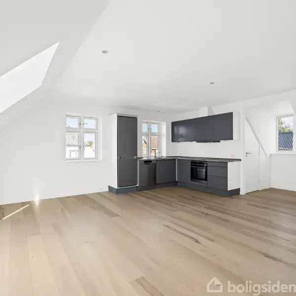 Et lyst, rummeligt køkken-alrum med grå skabe, indbygget ovn og komfur i et moderne design. Rummet har store lyse vinduer og skrå lofter med trægulve.