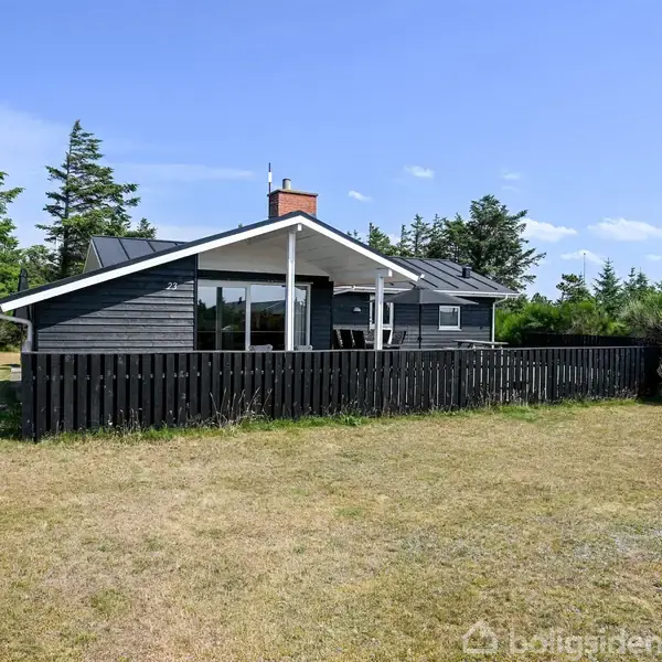 Et sortmalet sommerhus står stille omgivet af en græsplæne og træer under en blå himmel. Huset har en sort træterrasse indhegnet af et lavt stakit.