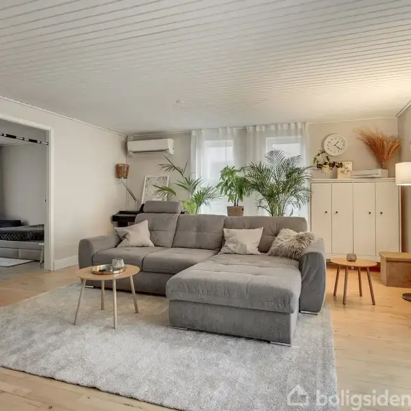 Grå sofa står på lys tæppe, omgivet af planter og små borde i en stue. Skab, ur, og gulvlampe i baggrunden. Bløde farver skaber en hyggelig atmosfære.