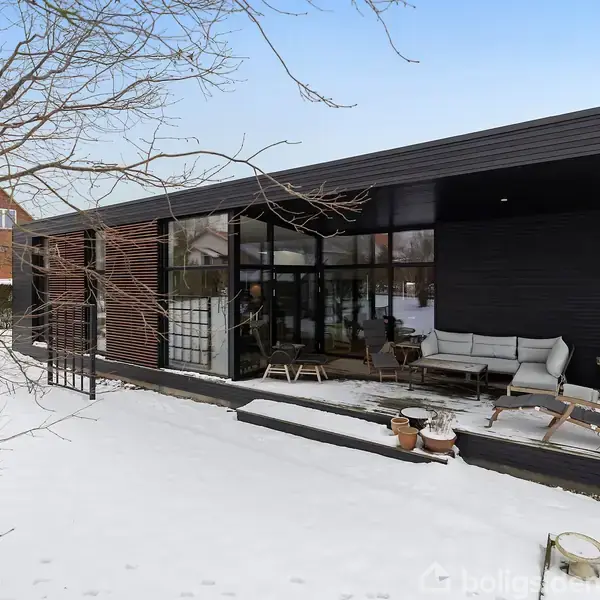 Et moderne hus med store glaspartier står stille i en snedækket have. Foran huset er en terrasse med møbler, omgivet af nøgne træer og en blå himmel.