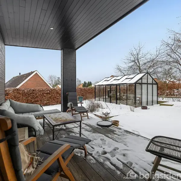 Overdækket terrasse med loungemøbler og bord; udsigt til snedækket have med drivhus i baggrunden og huse omgivet af buske.