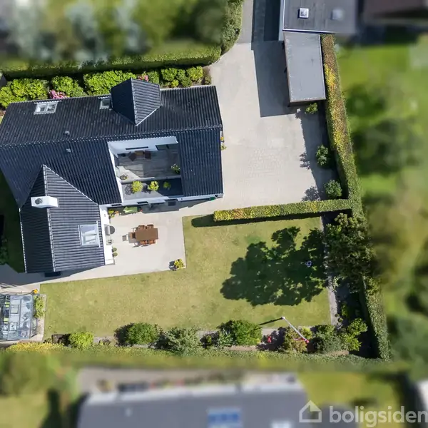 Et moderne, mørkt tagdækket hus står i centrum omkranset af en velplejet have med græsplæne og terrasse. Indkørsel og garage ses i baggrunden, omkranset af grønne hække.