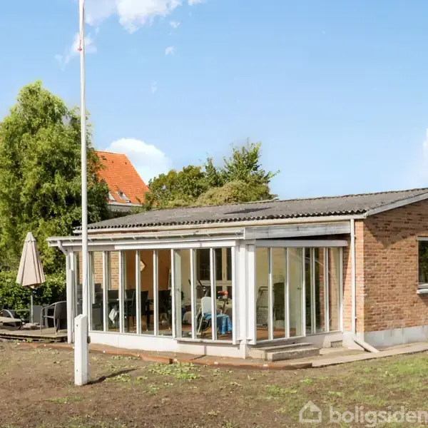 En murstensbygning med store vinduer er omgivet af en åben have med en flagstang og en parasol i nærheden. Bygningen står i et beboelsesområde med træer og andre huse.