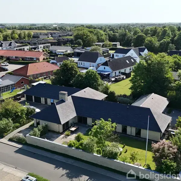 En moderne villa med sort tag står i en række af rækkehuse. Haven er omkranset af en grøn hæk. Baggrunden byder på flere huse og frodige træer i forstaden.