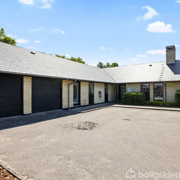 En moderne villa med fladt tag og lyse mursten står roligt. Den har garageporte og er omgivet af en asfalteret gårdsplads. Grønne træer ses i baggrunden.