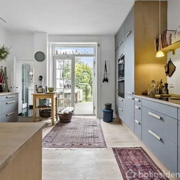 Køkken med blå skabe og træbordplader, oplyst af hængelamper. Krydderier og kogebøger arrangeret på hylder. Udgang til terrasse gennem glasdøre med udsigt til grøn have.
