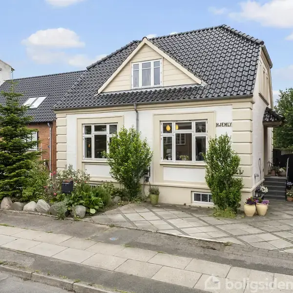 Et hvidt hus med en mørk, skråtstillet tag og kviste. Foran huset er der en have med buske og blomsterkrukker, flankeret af en fortovssti. Huset har teksten "HABENIK" ved indgangen.