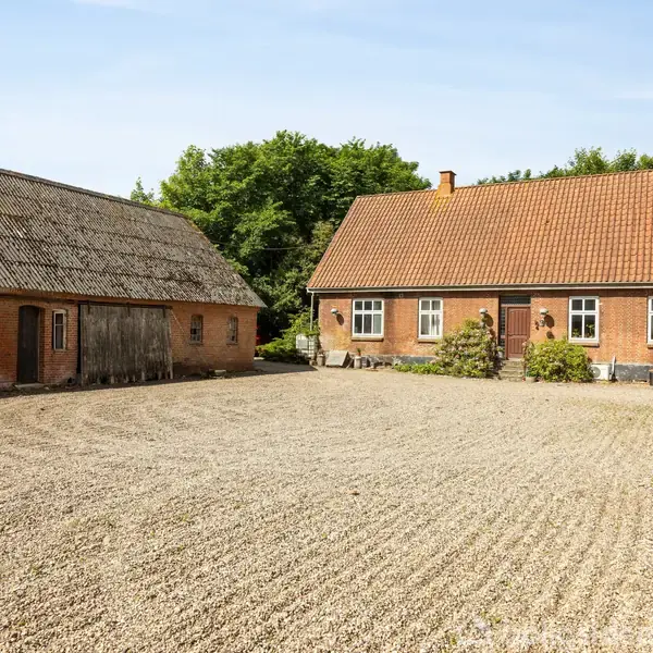 To bygninger står på en gårdsplads, den ene bruges som lade og den anden som bolig. En traktor er delvist synlig til højre. Grus dækker jorden, omgivet af træer.