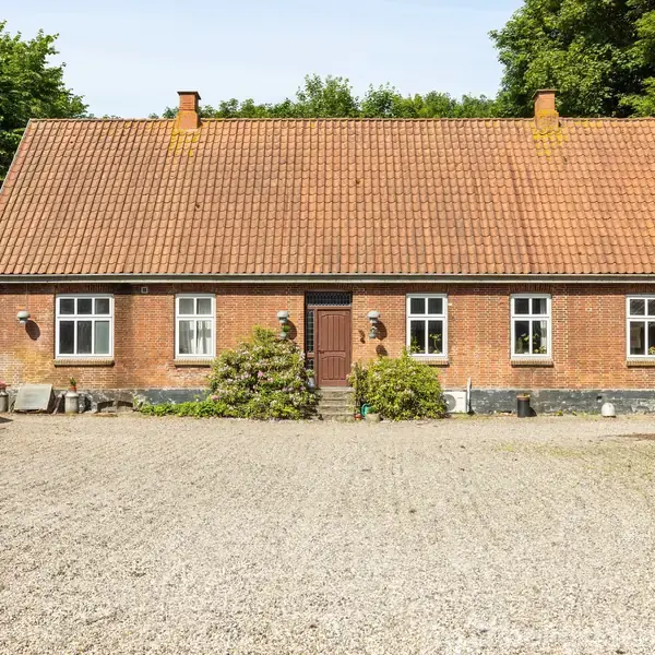 Rødt murstenshus med tre vinduer og et centralt placeret indgangsparti. Gruset gårdsplads foran og grøn bevoksning til venstre; omgivet af træer i baggrunden.