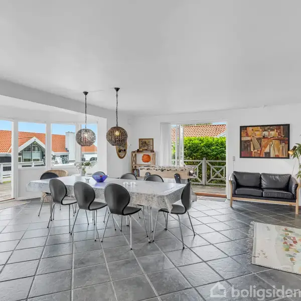 Spisestue med sort bord og stole, belyst af hængelamper. Store vinduer giver udsigt til røde taghuse. Sofa og plantereol står i hjørnet, hvide vægge omfavner rummet.