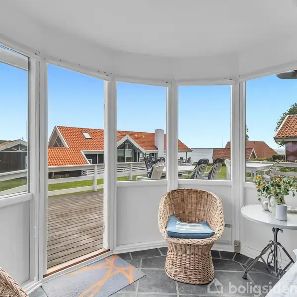 Rund kurvestol står stille i en lys udestue med udsigt over en terrasse og huse med røde tage. Et lille bord med en vase og stol supplerer rummet.