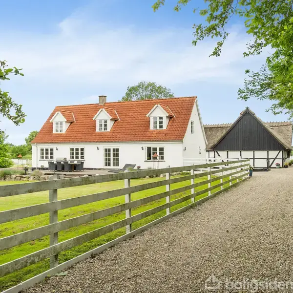 Et hvidt hus med rødt tegltag står stille langs en gruset vej. Ved siden af er en stor, sort-ladet skur og en grøn have med træer og hegn.
