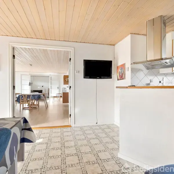 Et lyst køkken med flisegulv og hvide skabe. Et tv hænger på væggen. I baggrunden ses et spiseområde med bord og stole.