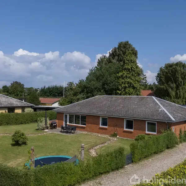 Et rødt murstenshus står omgivet af en velplejet have med en rund swimmingpool. En grussti løber langs hegnet, mens store træer og en blå himmel fylder baggrunden.