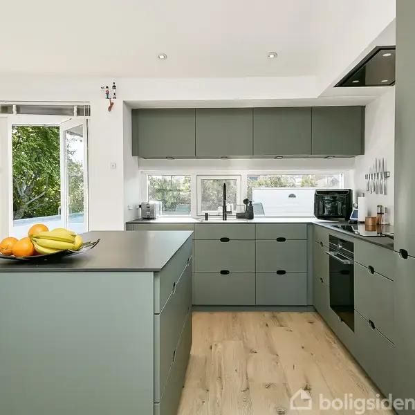 Et moderne køkken med grå skabe og en ø, hvor der ligger frugter. Der er store vinduer, der giver udsigt til en have. Rummet er lyst og luftigt.