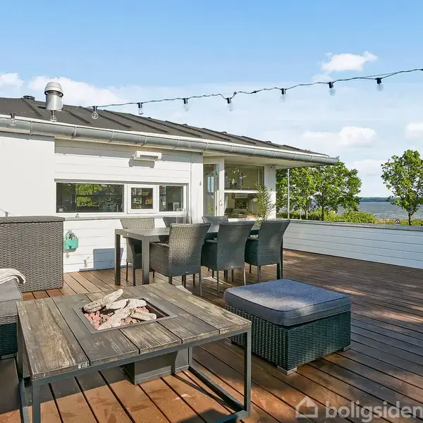 Terrasse med loungemøbler og spisebord, der skaber et hyggeligt udendørsrum. Bygningen i baggrunden har lyse vægge. Grønt landskab og vand ses i baggrunden under en blå himmel.