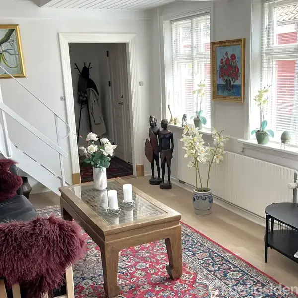 Stue med sofa og sofabord dekoreret med blomster og lys. Trappen fører opad. På væggen hænger malerier. Fjernsyn står på et lavt bord. Gulvtæppe med mønster på trægulv.