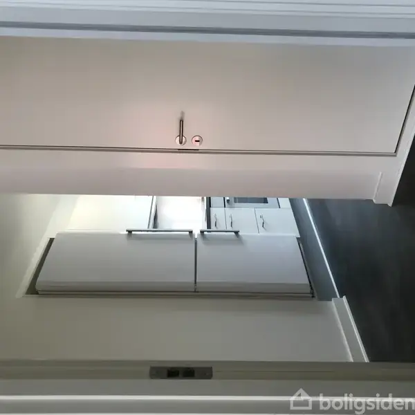 Hvidt køleskab står i et køkken med mørkt gulv, under en hvid skabsvæg. Køkkenet har minimalistisk indretning og er del af et større rum med hvide vægge.