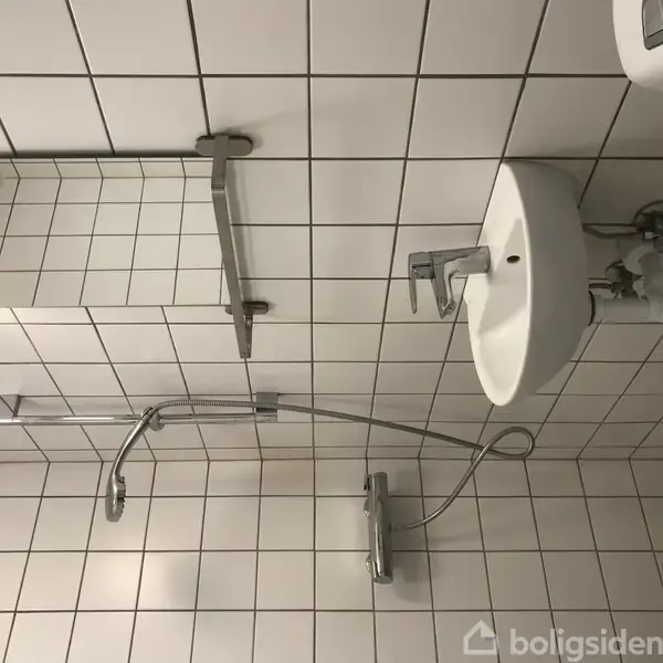 Håndvask med vandhane monteret på væggen over små hvide fliser. Spejl og lysarmatur hænger til venstre. Bruser og slange er til højre, også på flisevæg.