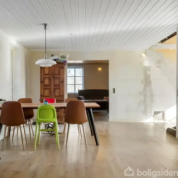 Spisebord med stole står centralt i et rum. En grøn barnestol er også til stede. Rummet har lyse trægulve, hvidt loft og en åben døråbning til et andet værelse med sofa.
