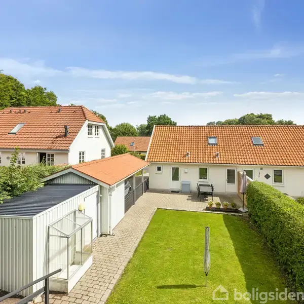 Et moderne hus med rødt tegltag står i en frodig have omgivet af grønne buske og træer. Foran huset ses en flisebelagt gang og en lille bygning til opbevaring.