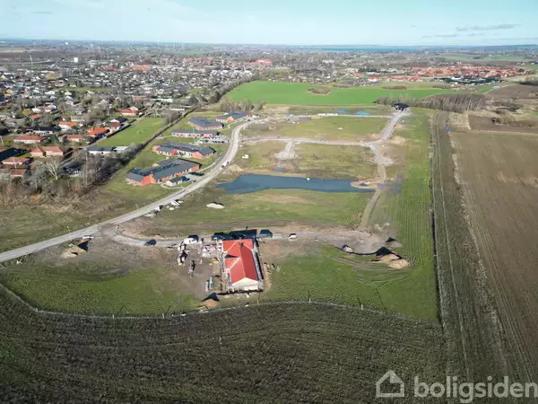 Bygning med rødt tag under konstruktion omgivet af grønne marker og små veje. I baggrunden ses en by med huse og åbne områder.