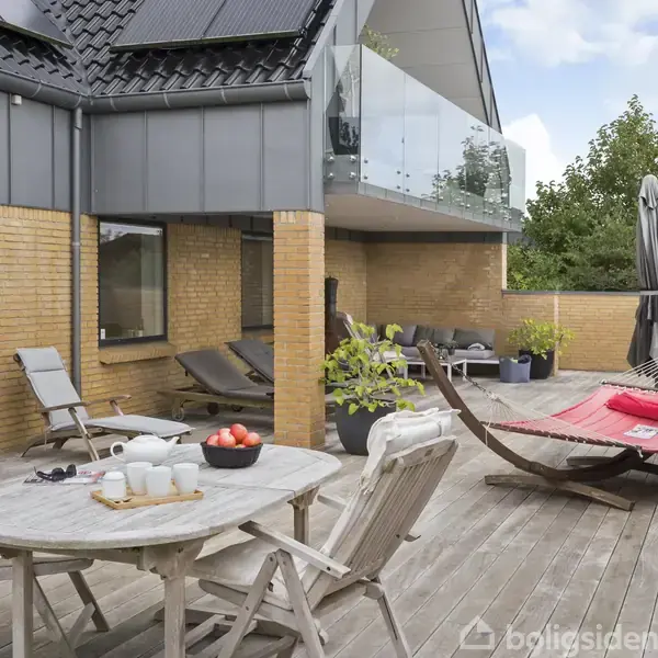 En træterrasse med et rundt bord og fire stole, en hængekøje samt liggestole. Terrassen er omgivet af murstensvægge og planter, under en blå himmel.