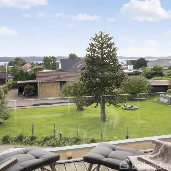 To liggestole står på en altan, med udsigt over en velplejet have med et stort, specielt træ. Området er beboelsesområde med huse i baggrunden, og himlen er let overskyet.