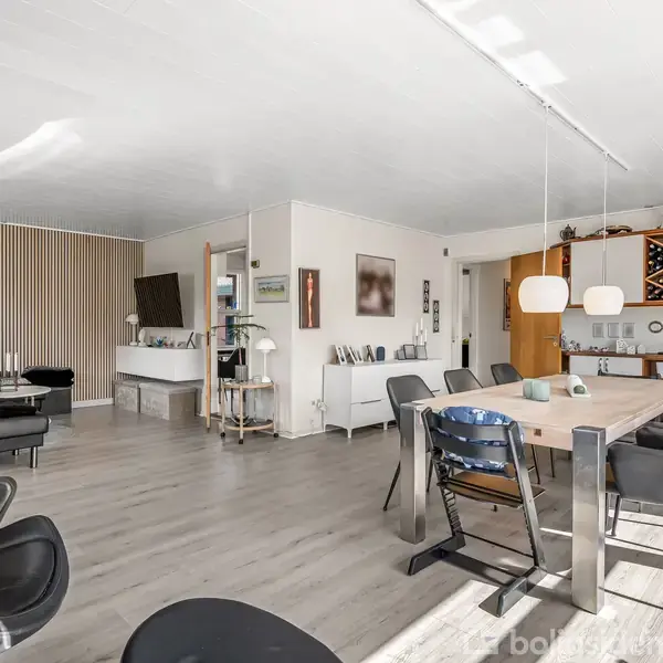 Stue med moderne møblering; et spisebord med stole og et opholdsområde med sorte lænestole. Trævæg, hylder og belysning skaber en hyggelig atmosfære. Masser af naturligt lys.