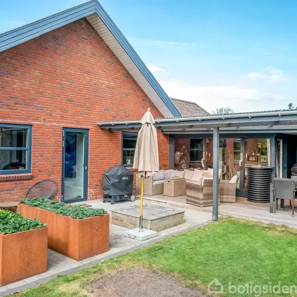 Rødt murstenshus med en overdækket terrasse. To store plantekasser står i forgrunden. Terrassen har en parasol, grill og møbler, omgivet af en grøn have.