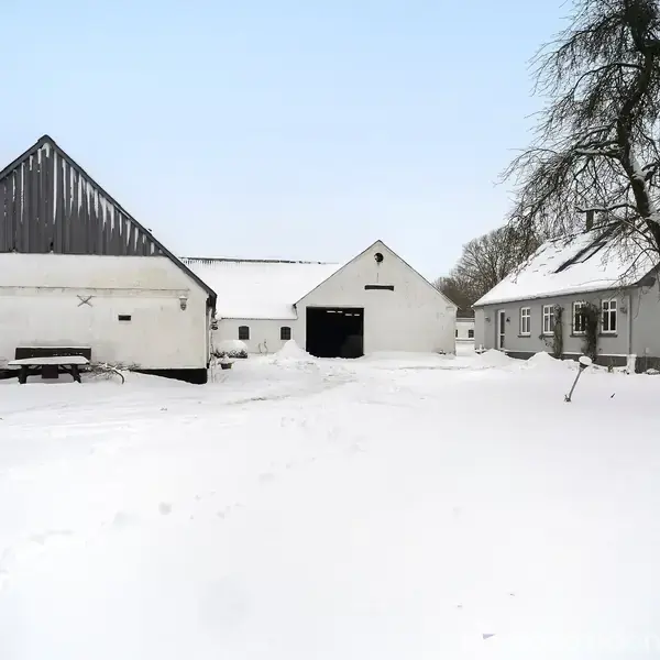 Et gårdanlæg dækket af sne viser tre hvide bygninger med grå tage. En gårdsplads med et sneklædt træ og en bænk skaber en vinterlig atmosfære.