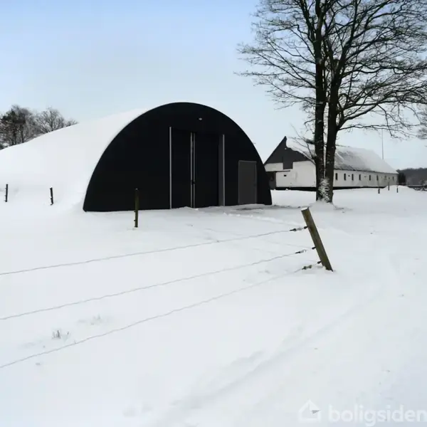 Et sort halvcirkelformet skur står stille i et snedækket landskab ved siden af et træ og et hegn. I baggrunden ses en lang, hvid bygning.