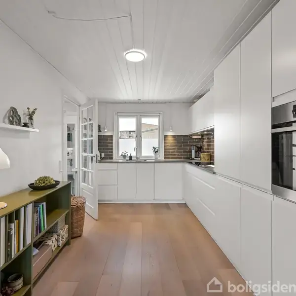 Et moderne køkken, hvor indbyggede hvide skabe og en ovn dominerer rummet. En grøn reol står til venstre med bøger og pynteting. Et vindue giver naturligt lys.