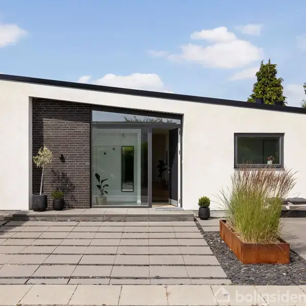 En moderne, hvid bungalow med fladt tag har store vinduer og en hoveddør. Foran huset er en flisebelagt terrasse med planter i krukker. Omgivet af hække og træer.