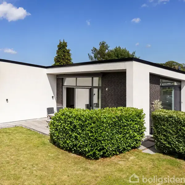 En moderne villa med fladt tag står i en frodig have. Store vinduer reflekterer dagslys, omgivet af velplejet græsplæne og buske. Klar himmel med lette skyer i baggrunden.