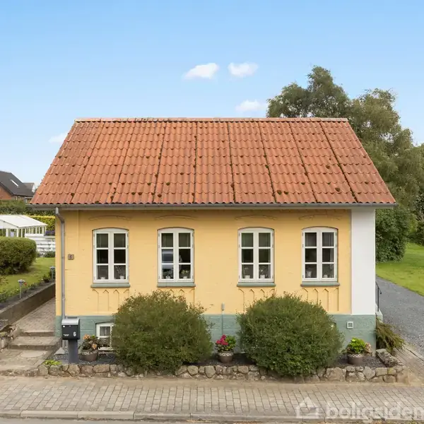 Et lille, gult hus står stille ved en brolagt vej. Det har rødt tegltag, tre buede vinduer og omgives af grønne buske og græsplæne med en gravet vej ved siden.