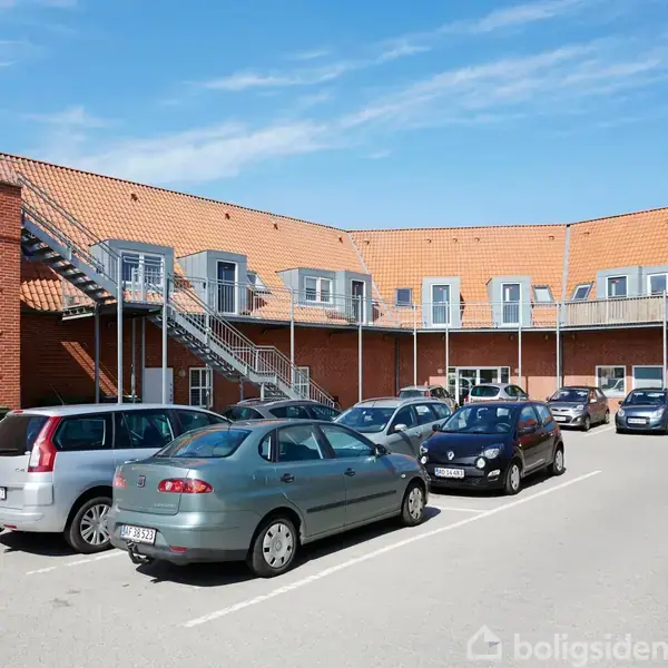 Biler holder parkeret foran en toetagers bygning med røde mursten og orange tegltag. Udendørs trapper giver adgang til øverste etage. Klar blå himmel i baggrunden.