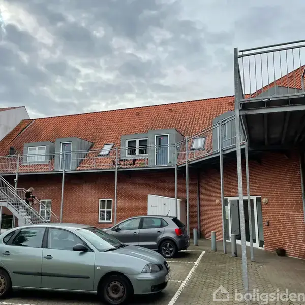 Biler står parkeret foran en murstensbygning med rødt tegltag og udvendige trapper til første sal, under en overskyet himmel.