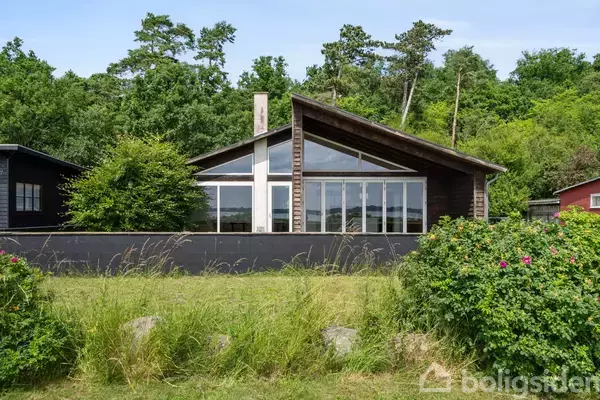 Sommerhus med store vinduespartier står stille i en grøn have omkranset af buske og træer. Bagved ses en skov, hvilket skaber en fredelig og naturlig atmosfære.