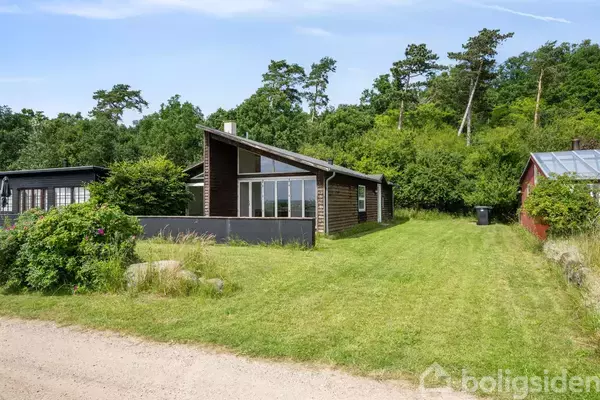 Et moderne sommerhus med store vinduer står på en velplejet græsplæne omgivet af buske og træer i en skovbevokset område.