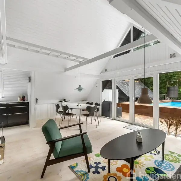 Stue med kamin, grå stol og sort sofabord står på et farverigt tæppe. Rum med åbent højt loft. Store vinduer åbner ud til en terrasse med pool og skov i baggrunden.